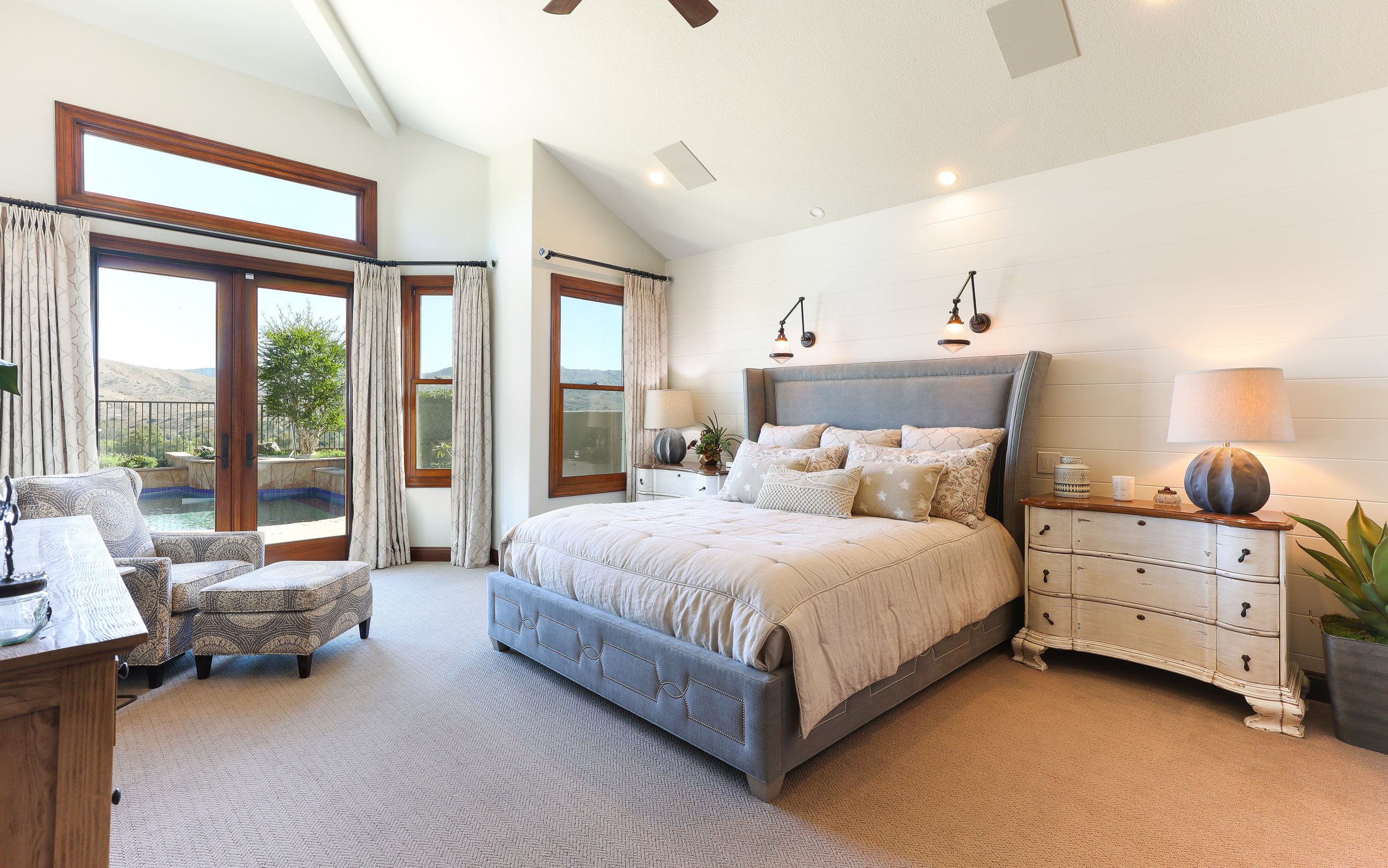 A bedroom with a large bed, nightstands, and lamps, with windows showing an outdoor view of hills and trees.