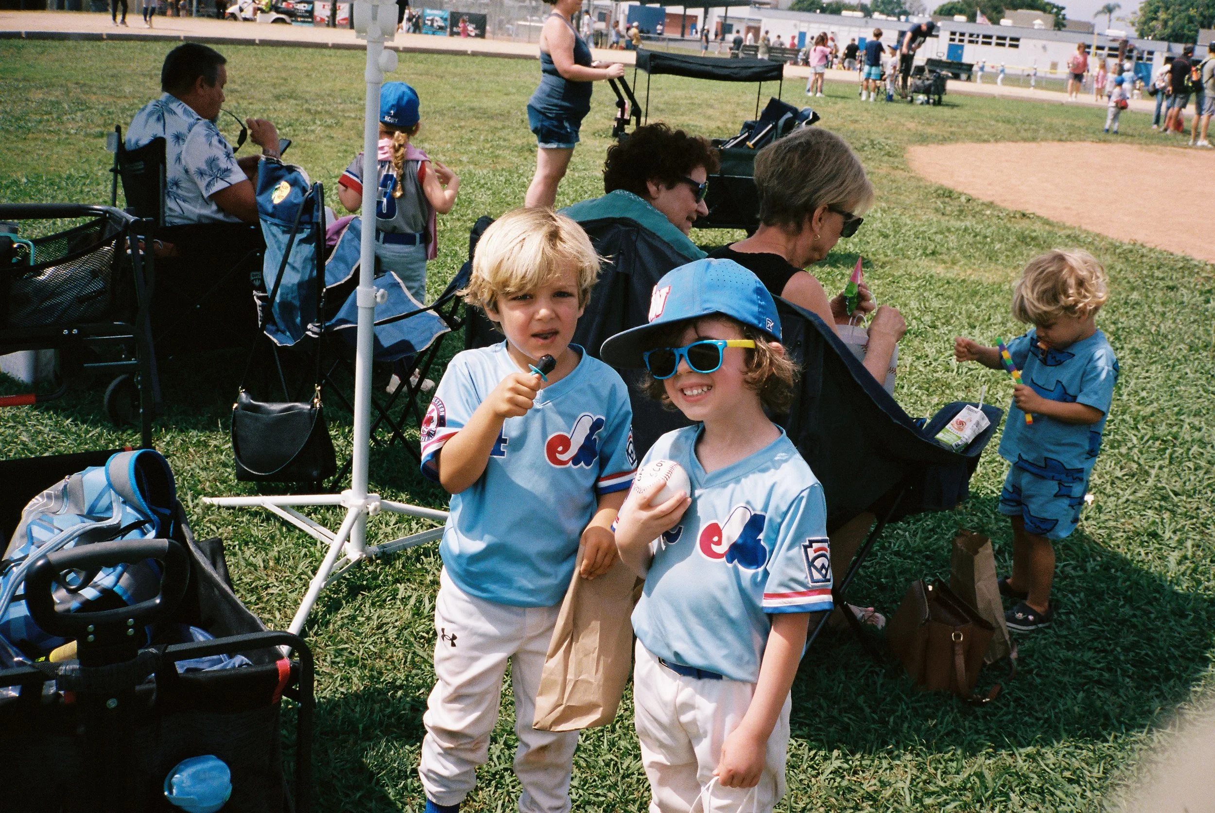 Group of children and adults sitting and standing on grass at a park during a baseball game, with some children in uniform holding baseball gloves and a ball, and a baseball field with spectators in the background.