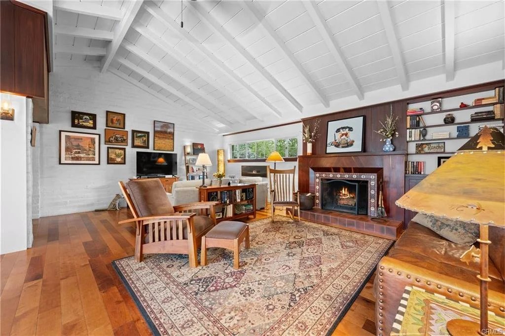 Living room with white vaulted ceiling, hardwood floors, a brick fireplace with a wooden mantel, a patterned area rug, leather and upholstered chairs, a wooden bookcase, and framed artwork on the wall.