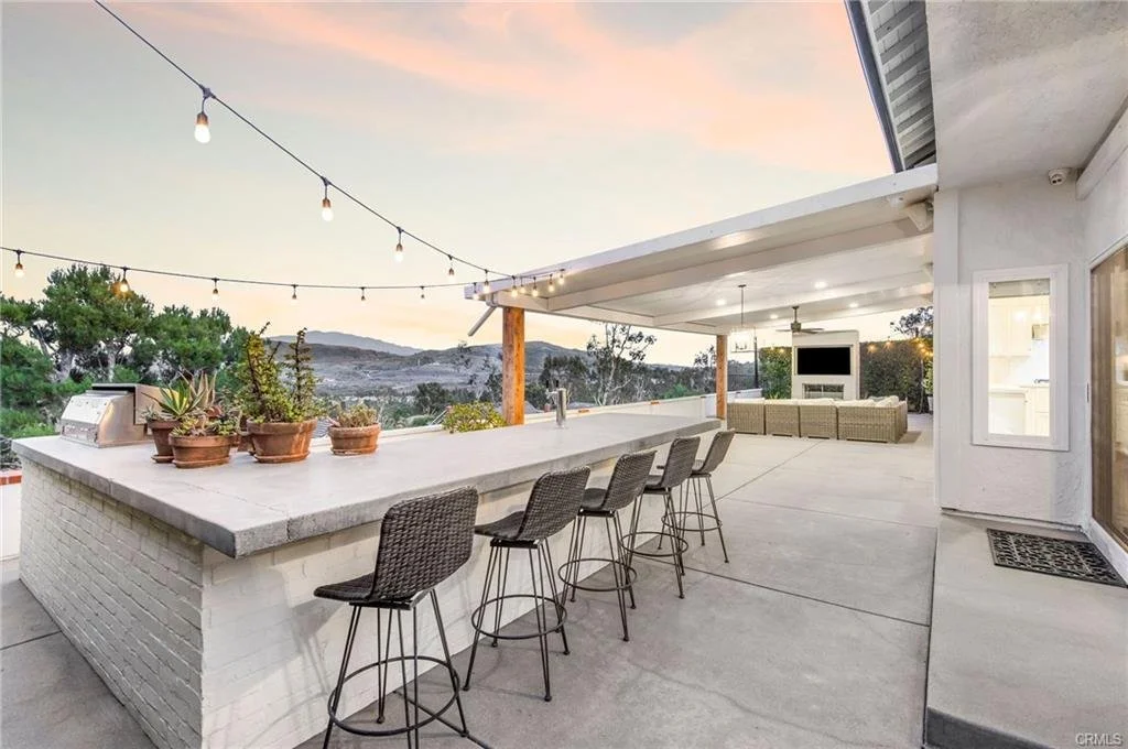 Outdoor patio with string lights, a bar with bar stools, potted plants, a sitting area with a television, and scenic mountain views at sunset.