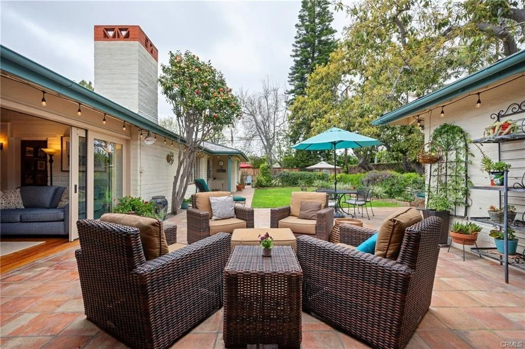 Outdoor patio with wicker furniture, potted plants, a blue patio umbrella, and a lawn area with trees and shrubs.