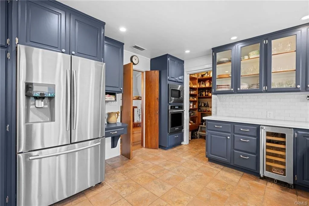 Kitchen with blue cabinets, stainless steel refrigerator, built-in microwave and oven, tile floor, open door to pantry.