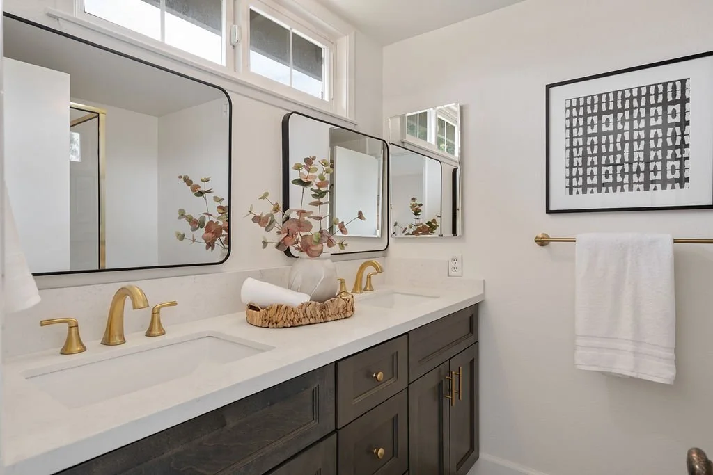 Modern bathroom with dark cabinetry, white countertop, gold fixtures, three mirrors, and a framed black-and-white abstract art piece