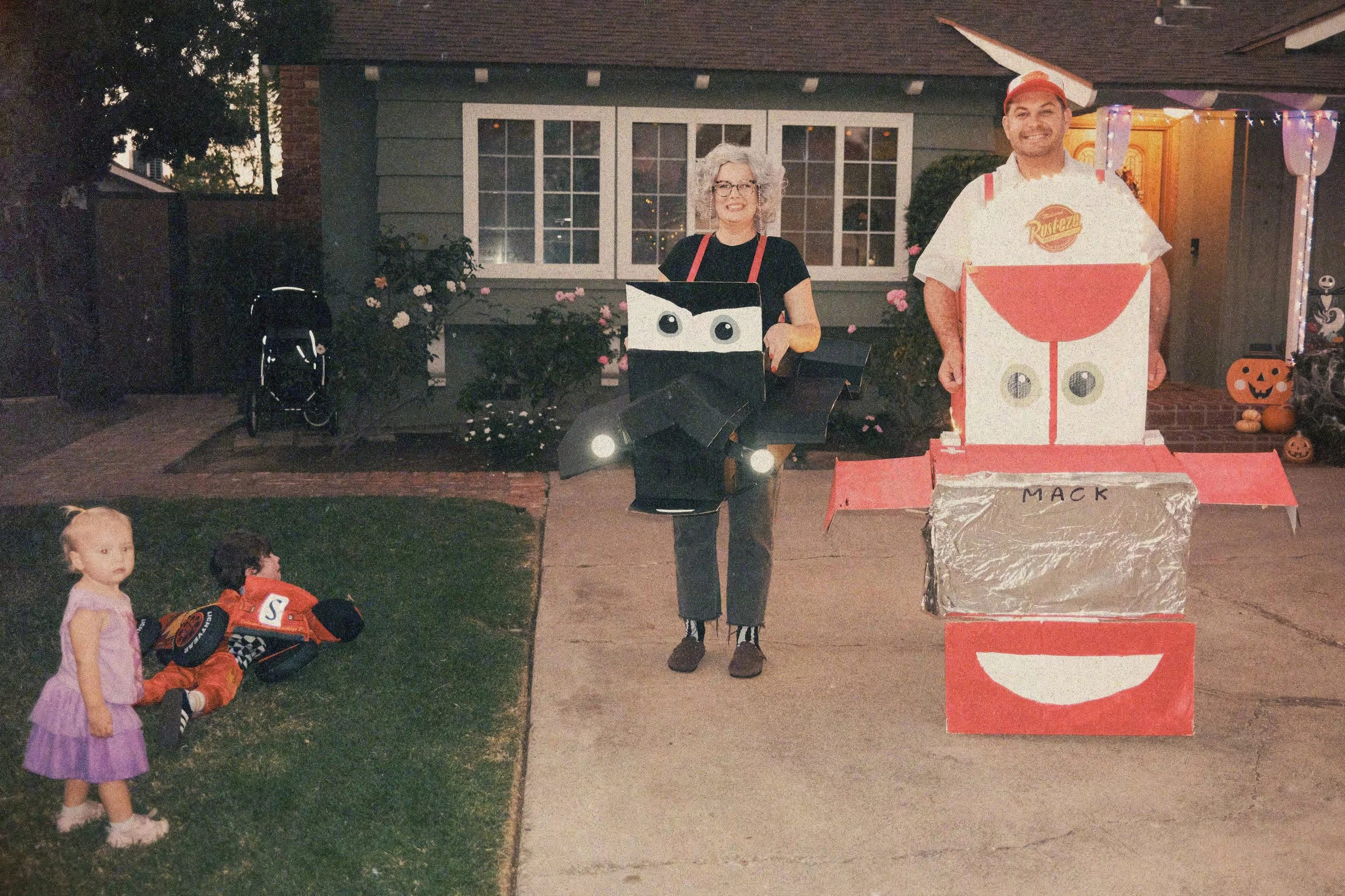 Two smiling adults dressed as cartoon characters with large boxes decorated as a face and a cup, standing outdoors in front of a house during Halloween. Two young children are nearby, with one girl in a purple dress and another child in a firefighter costume sitting on the grass. Halloween decorations include pumpkins and a pumpkin-shaped ornament.