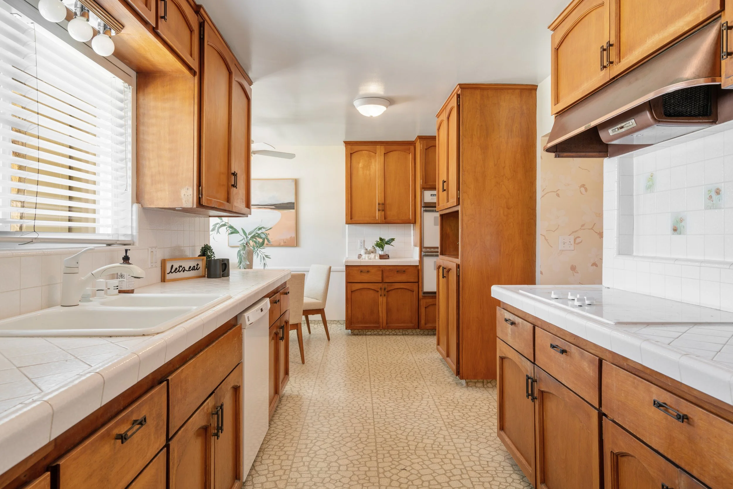 Kitchen with wooden cabinets, tiled countertops, white sink, window with blinds, and a white stove, with pattern linoleum floor.