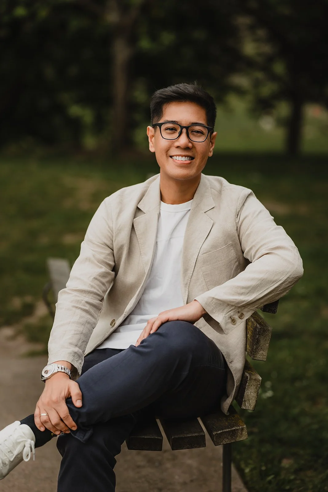 A young man with glasses, wearing a beige blazer and white shirt, sitting on a park bench and smiling.