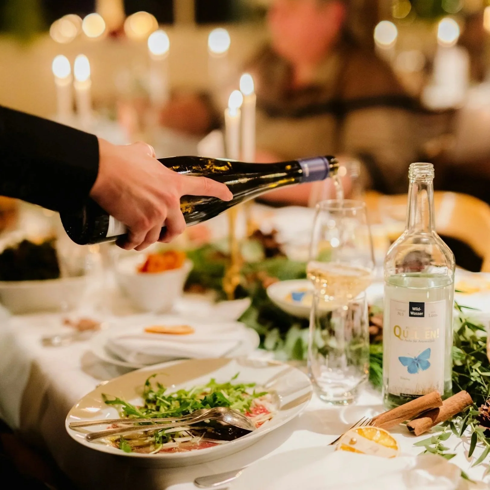 Person pouring drinks into glass at a festive dinner table with food, candles, and decorations visible.