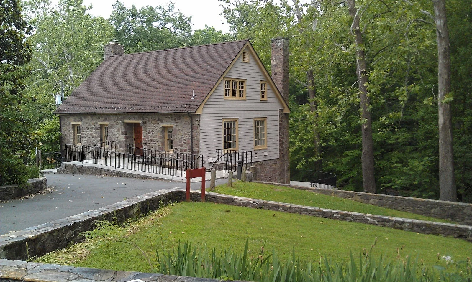 A house with stone and wood siding, surrounded by trees and a grassy lawn, with a paved driveway and a red sign near the entrance.