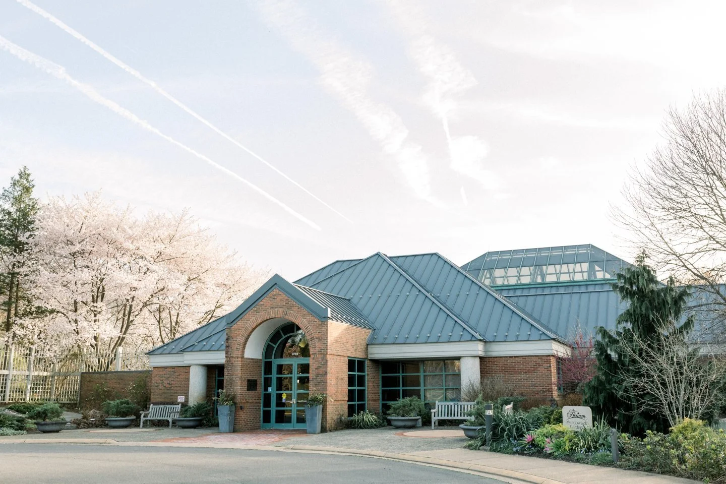 A brick and glass building with a blue metal roof, surrounded by trees and flowers, under a sky with contrails.