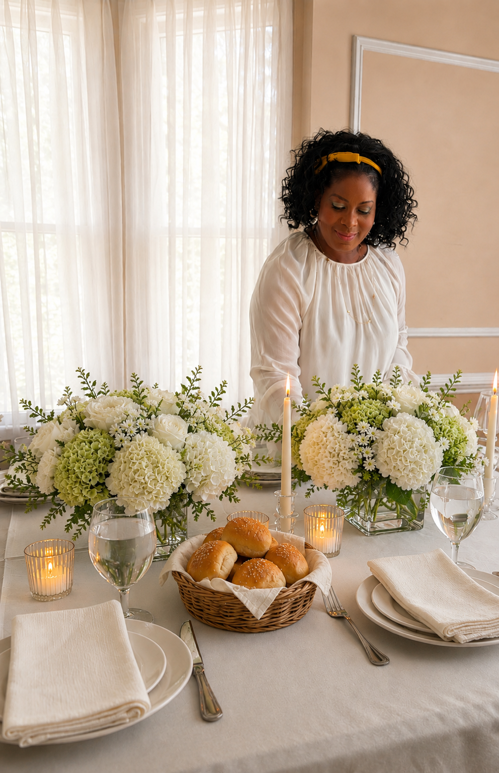 Woman setting a formal dining table with white flowers, candles, bread rolls, and glasses of water.