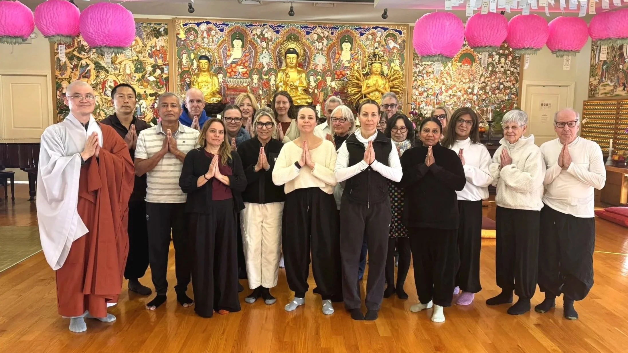 Group of diverse people standing in a Buddhist temple, praying with hands together in front of richly decorated altar with golden Buddha statues, intricate murals, pink paper lanterns hanging from ceiling, and wooden floor.