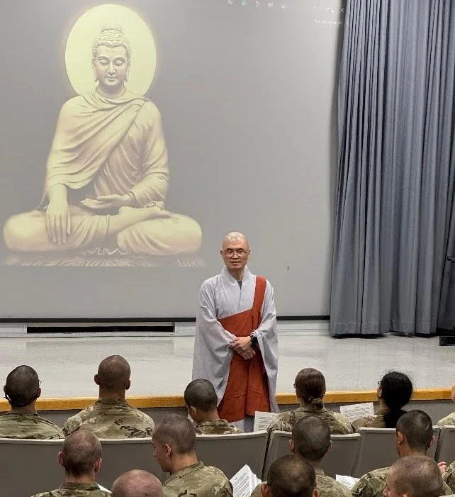 A monk standing in front of a group of soldiers during a presentation or lecture. In the background, there is a large projection of a Buddha statue with a halo.