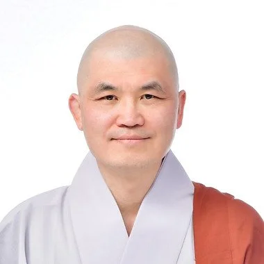 A smiling man with a shaved head wearing traditional white and brown Buddhist robes
