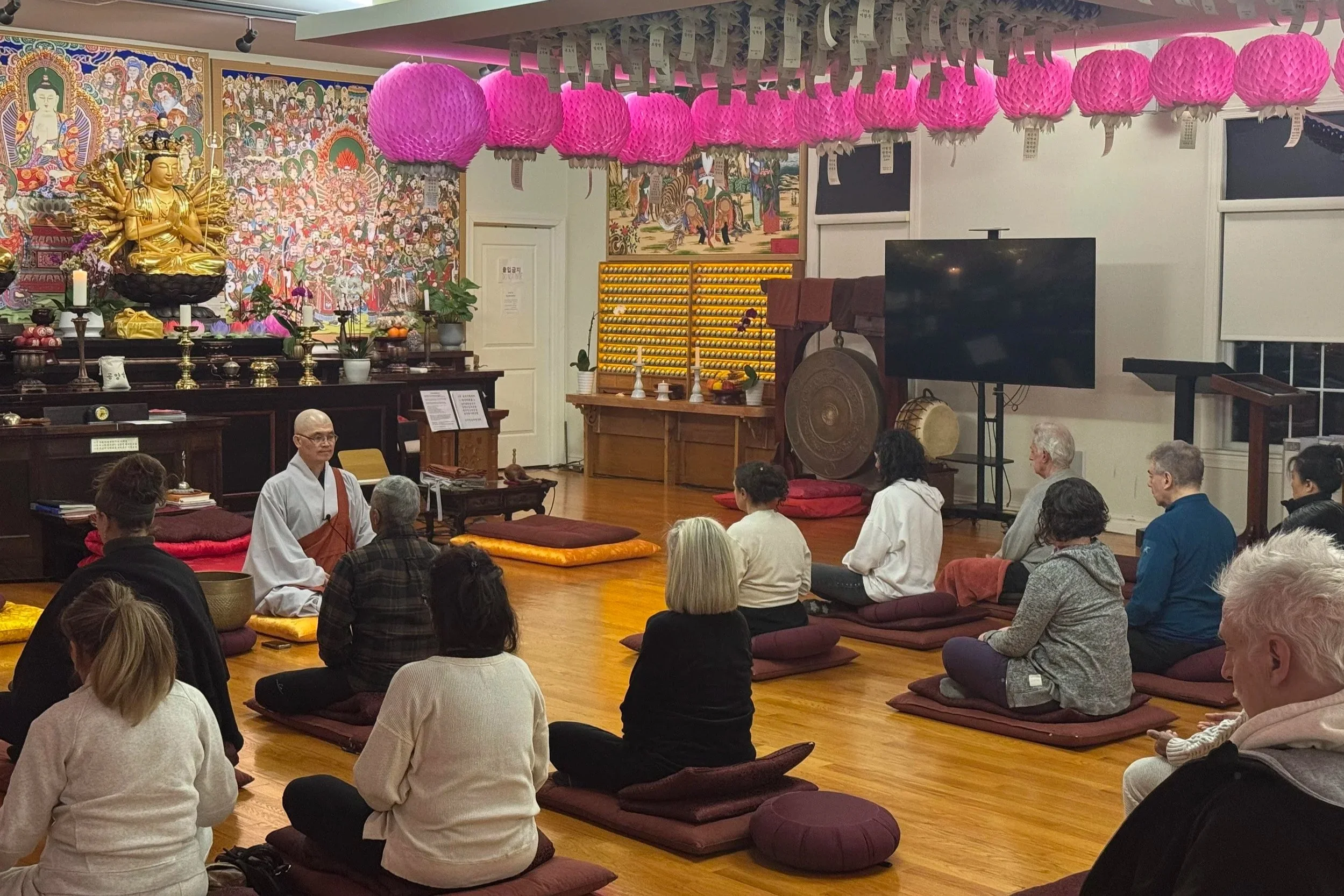 People sitting on cushions in a meditation session inside a Buddhist temple, with a monk leading the session. The temple features pink paper lanterns hanging from the ceiling, a large golden statue of a Buddha, and colorful traditional artwork on the walls.
