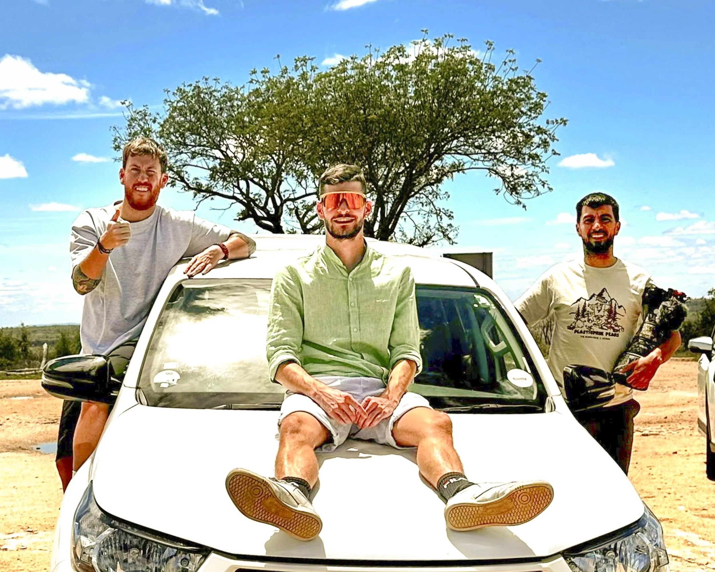 Tre uomini sorridenti su un'auto bianca all'aperto sotto un albero, con cielo blu e nuvole sparse.