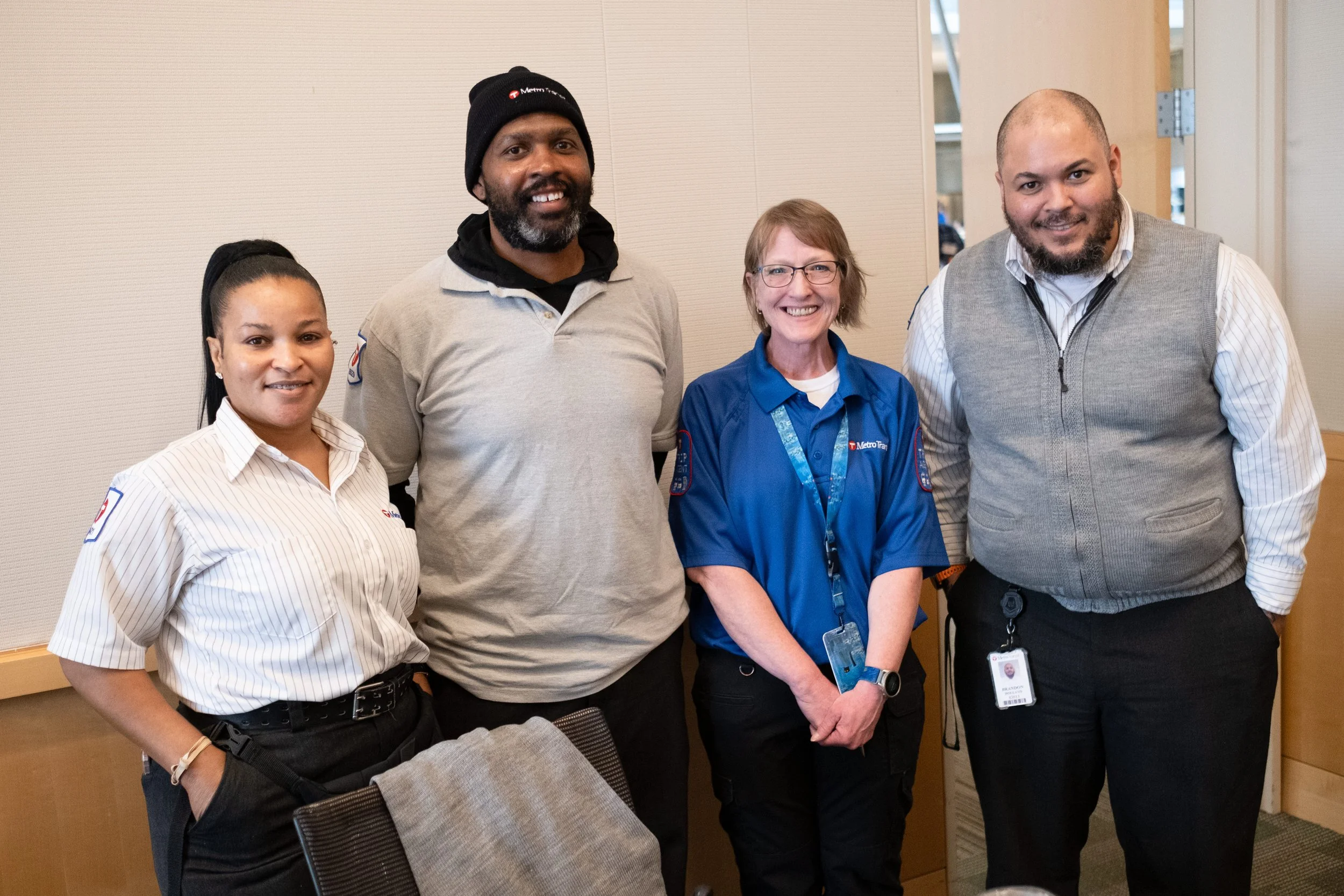 Group of four diverse adults, standing together indoors, smiling at the camera.