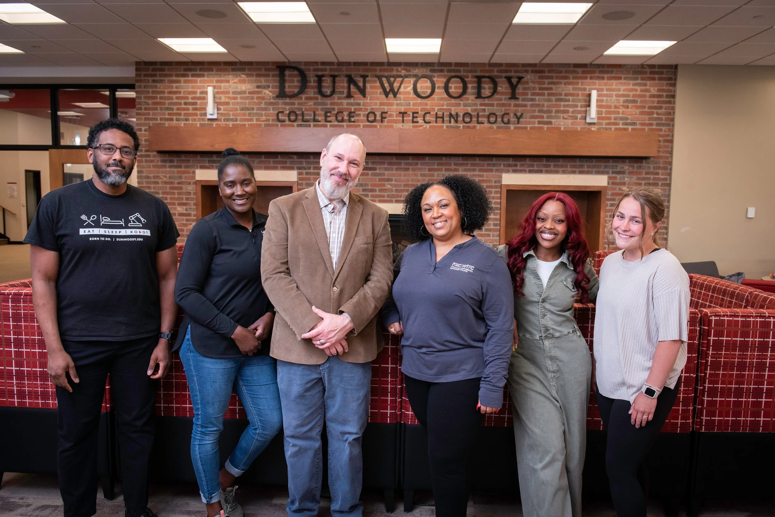 A group of six diverse people standing together inside a building with a brick wall and the sign 'DUNWOODY COLLEGE OF TECHNOLOGY' behind them. They are smiling and appear to be in a professional or academic setting.
