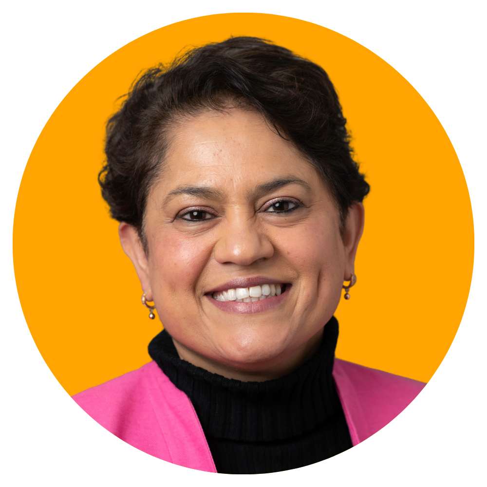 A woman with short dark hair, wearing a pink blazer, black turtleneck, and earrings, smiling against a solid yellow background.