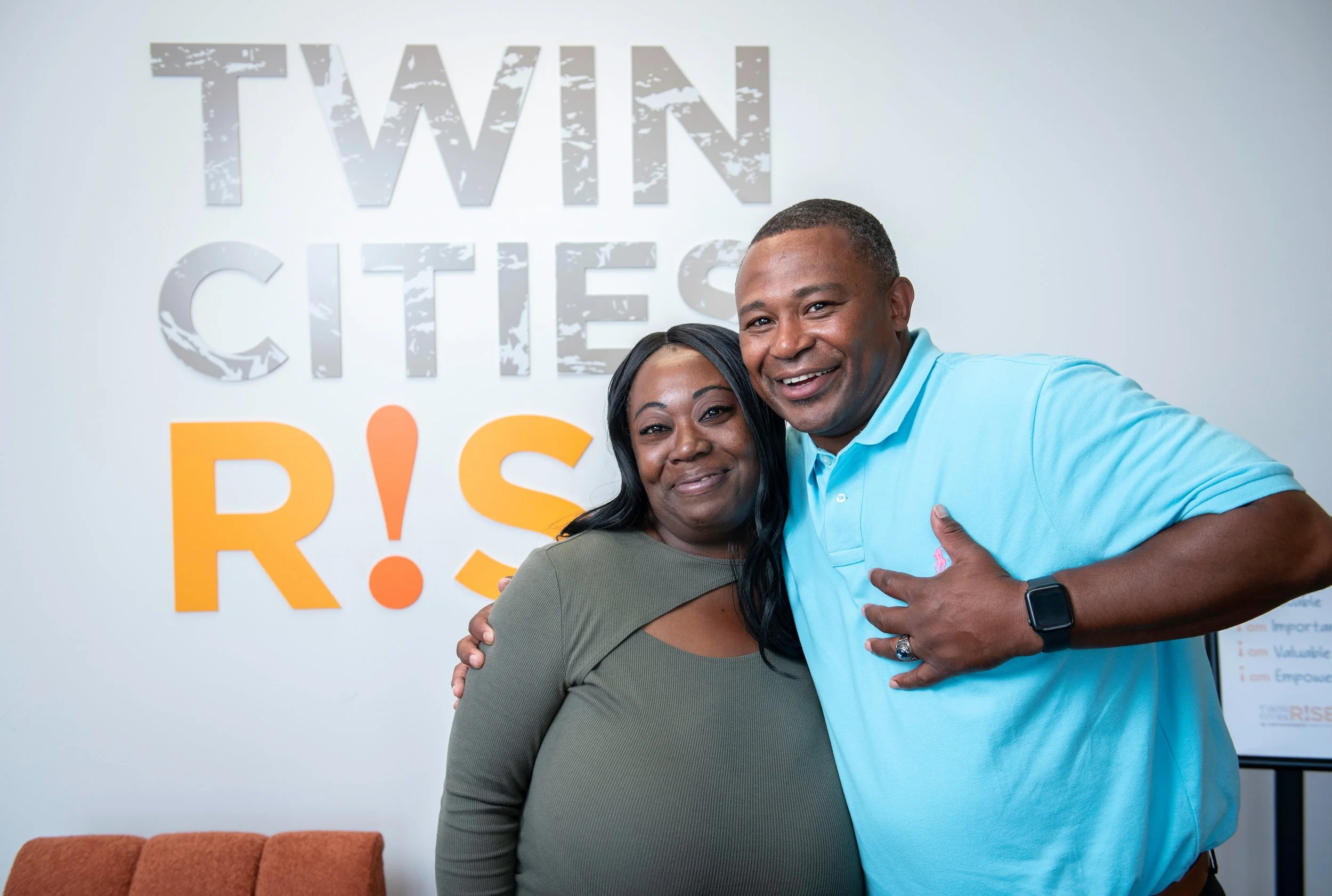 Two smiling people hugging in front of a wall with the words 'Twin City RISE'.
