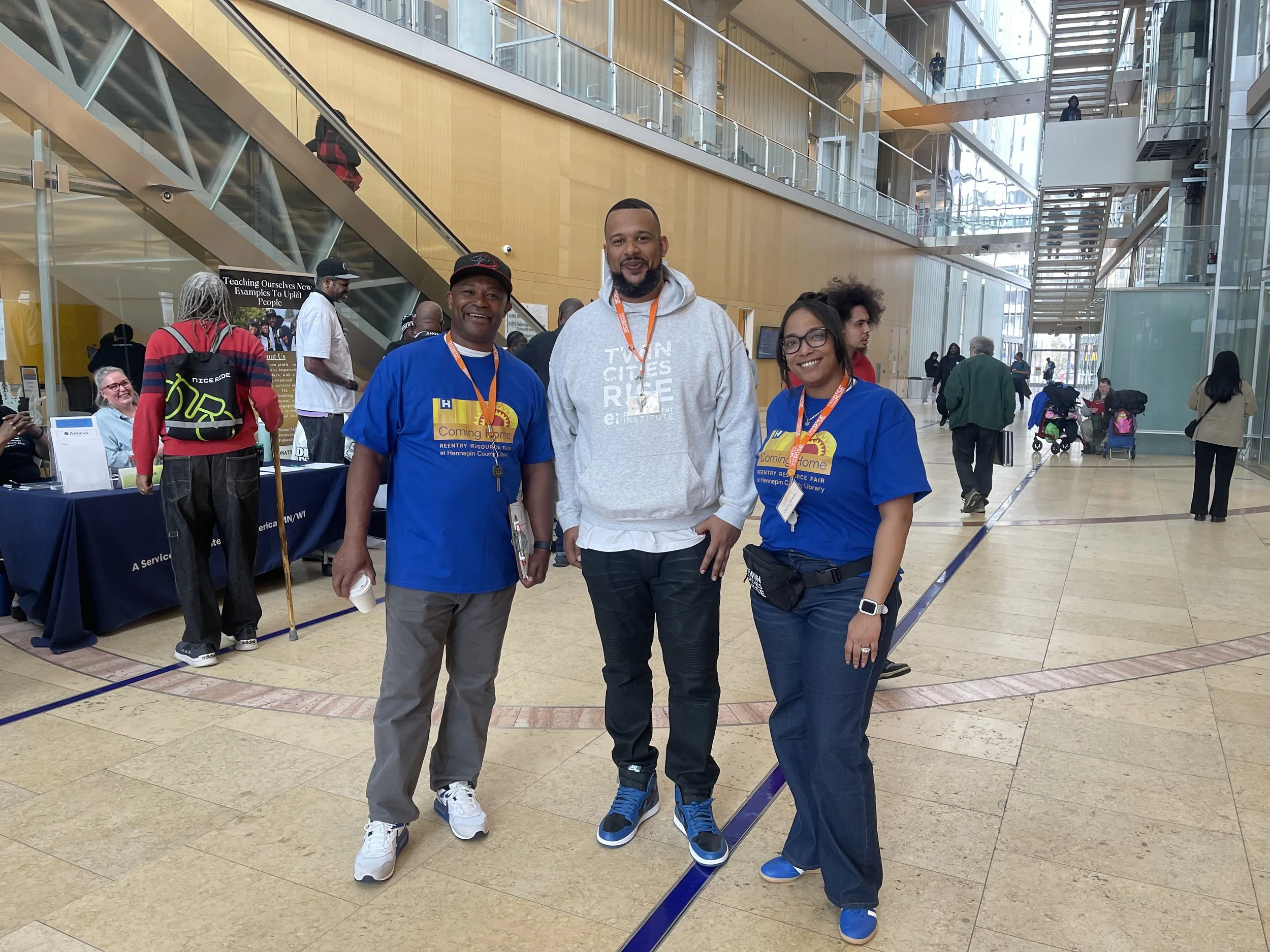 three people standing together in a modern indoor atrium with glass walls and multiple floors. The two on the left are men wearing blue shirts, and the person on the right is a woman in a blue shirt. They are all smiling at the camera, and there are other people and tables in the background, suggesting a busy event or conference.