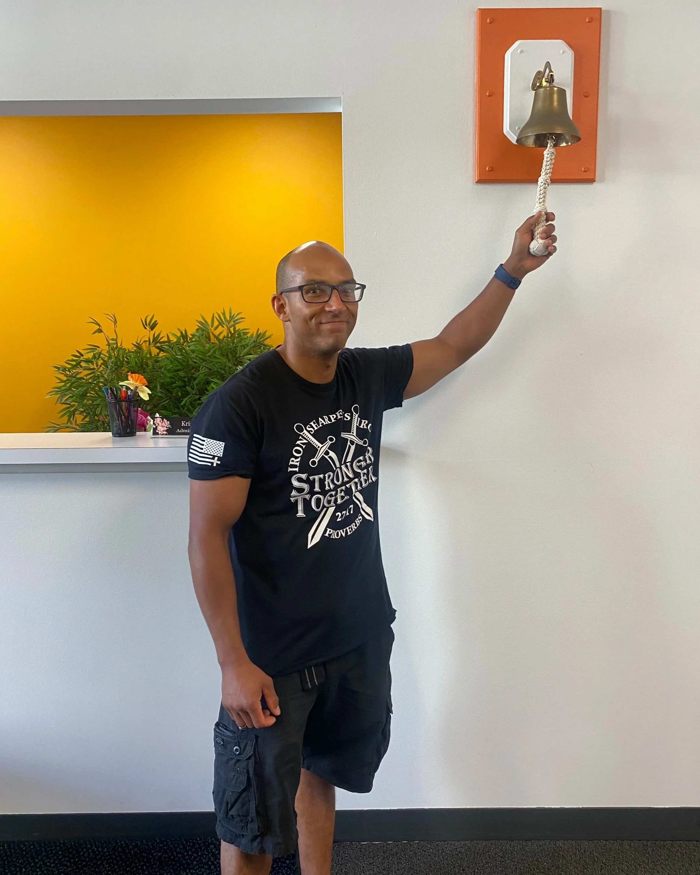 A man in a black t-shirt and shorts standing inside an office, ringing a brass bell mounted on a white wall with orange border. There is a yellow wall in the background.