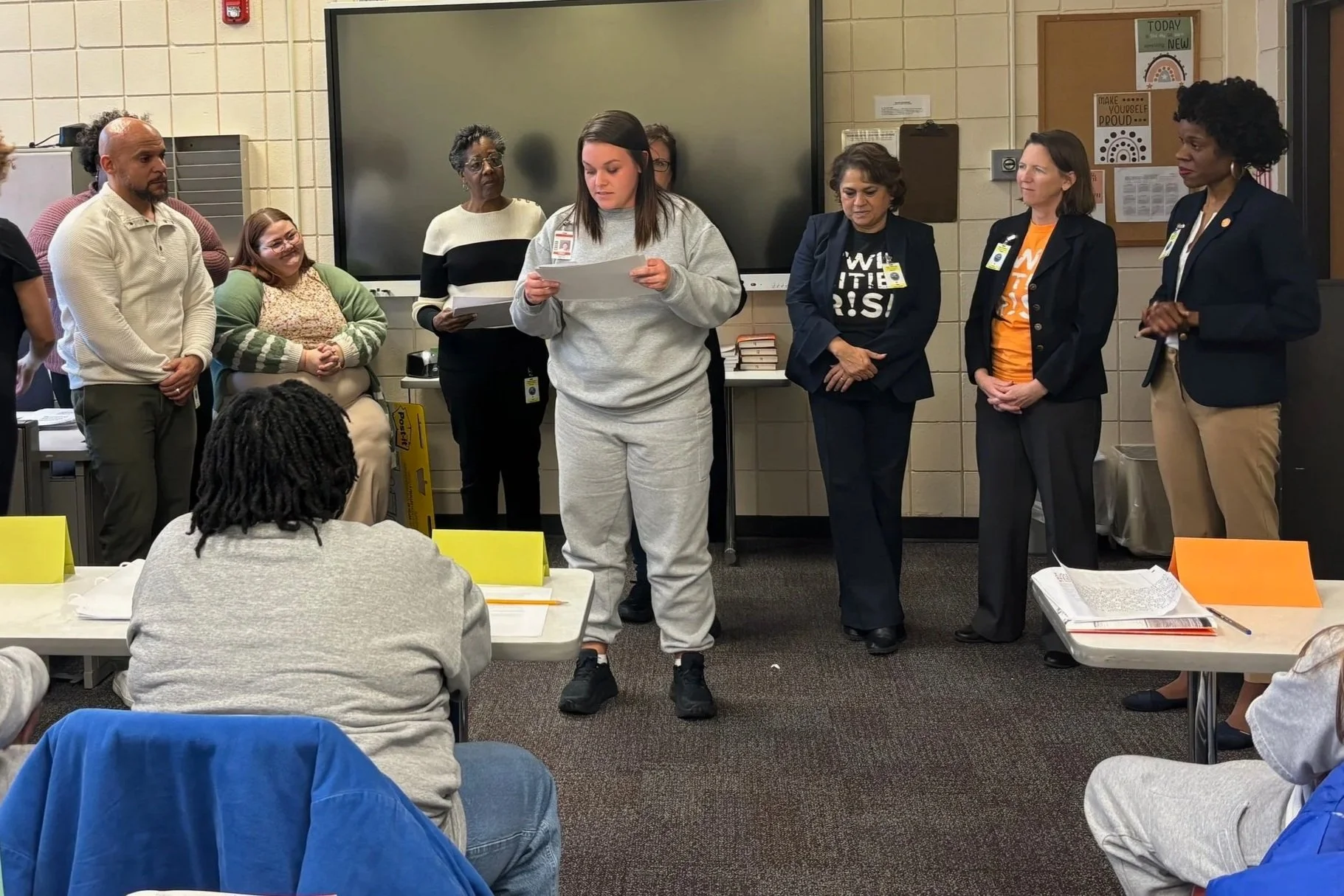 A group of diverse adults standing and sitting in a classroom or meeting room. One woman in casual attire appears to be reading or presenting, while others listen attentively. The room has educational posters and a large digital board on the wall.
