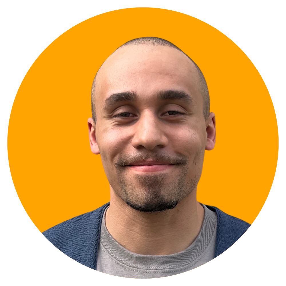 A smiling man with a closely shaved head and a light goatee, wearing a gray shirt and navy blazer, against an orange background.