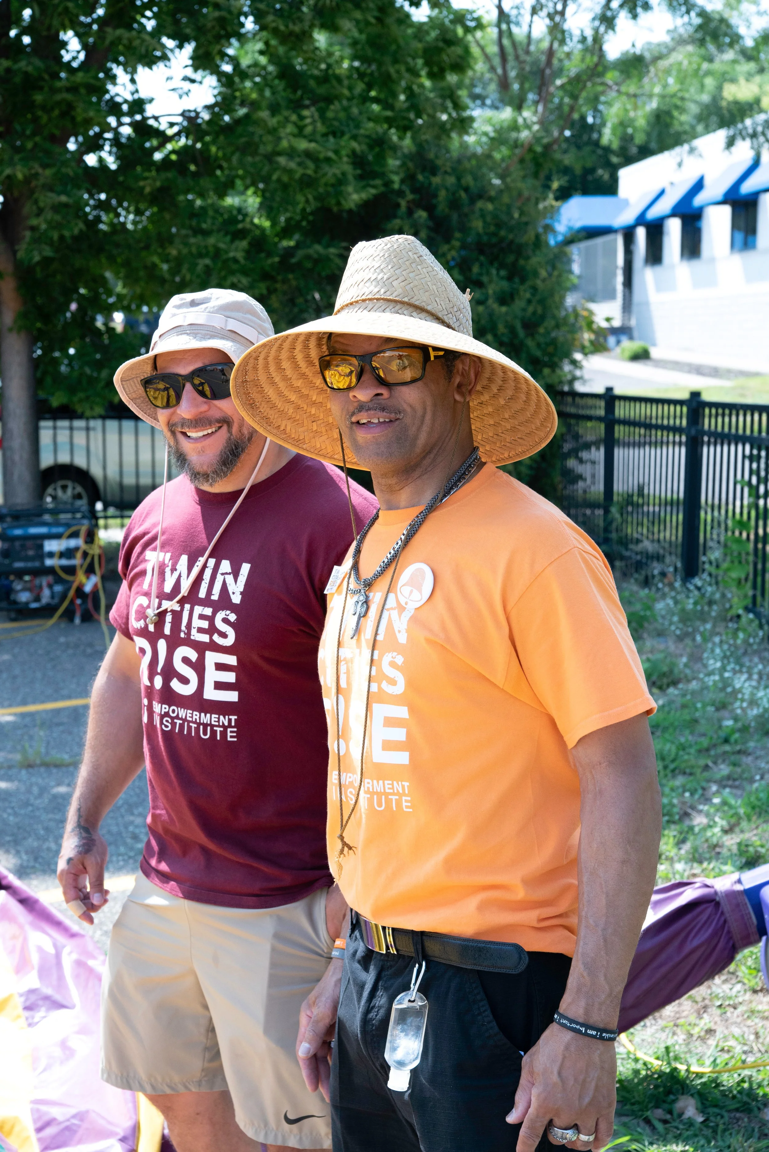 Two men outdoors wearing sunglasses and hats, one in a maroon T-shirt and the other in an orange T-shirt, smiling and walking together.