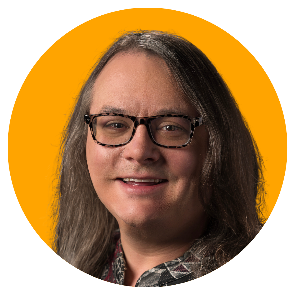 Close-up portrait of a person with long, wavy hair and glasses, smiling in front of a bright orange background.