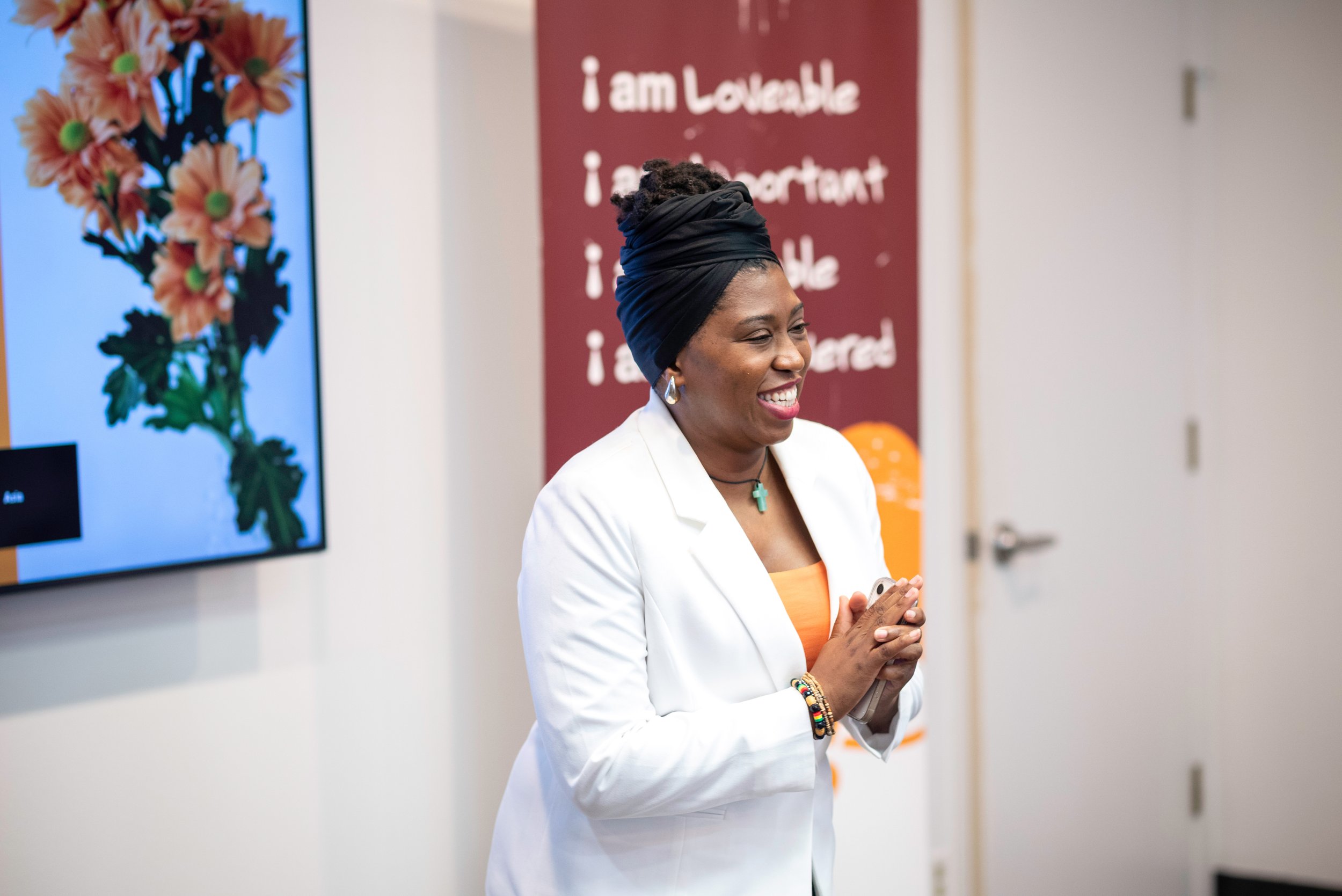A woman with dark hair wrapped in a black headscarf, dressed in a white blazer and orange top, smiling while holding a smartphone in a room with a large screen displaying flowers and a red wall with white text.