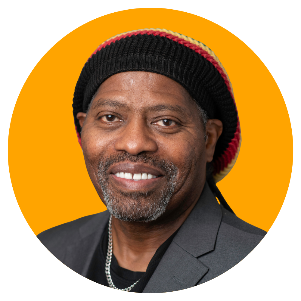 Close-up of a smiling African American man wearing a beanie with red, yellow, and black stripes, a necklace, and a dark blazer against an orange background.