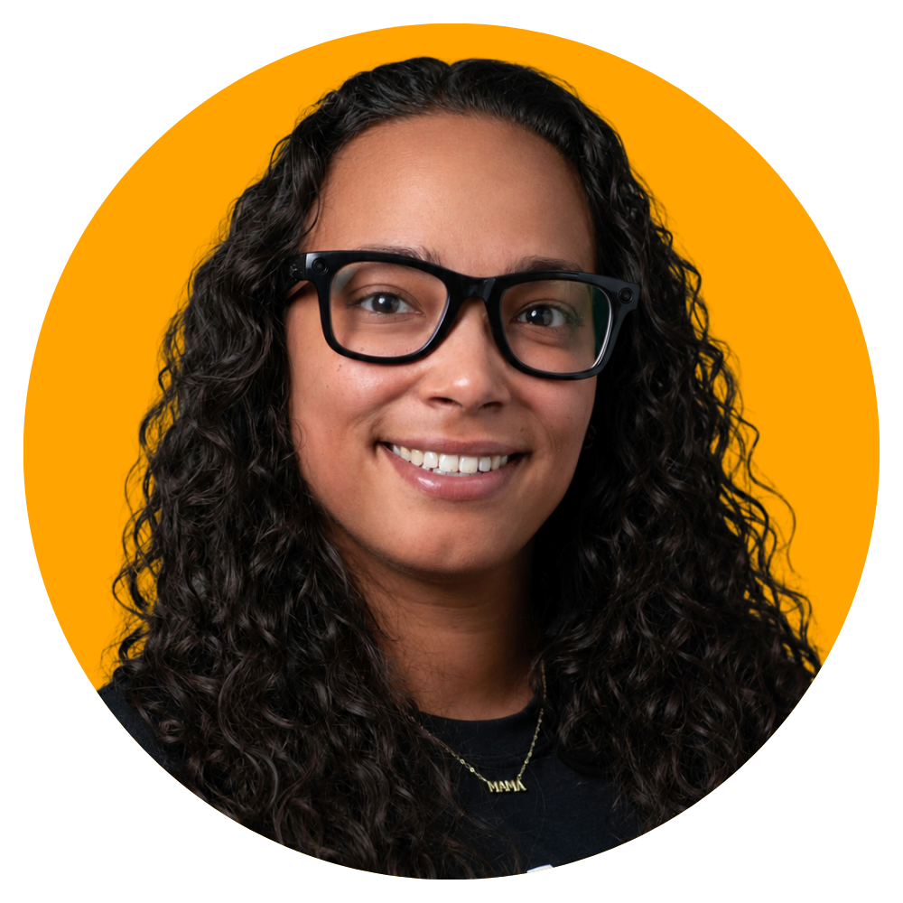 A woman with curly hair wearing black glasses and a black top, smiling against a yellow background.