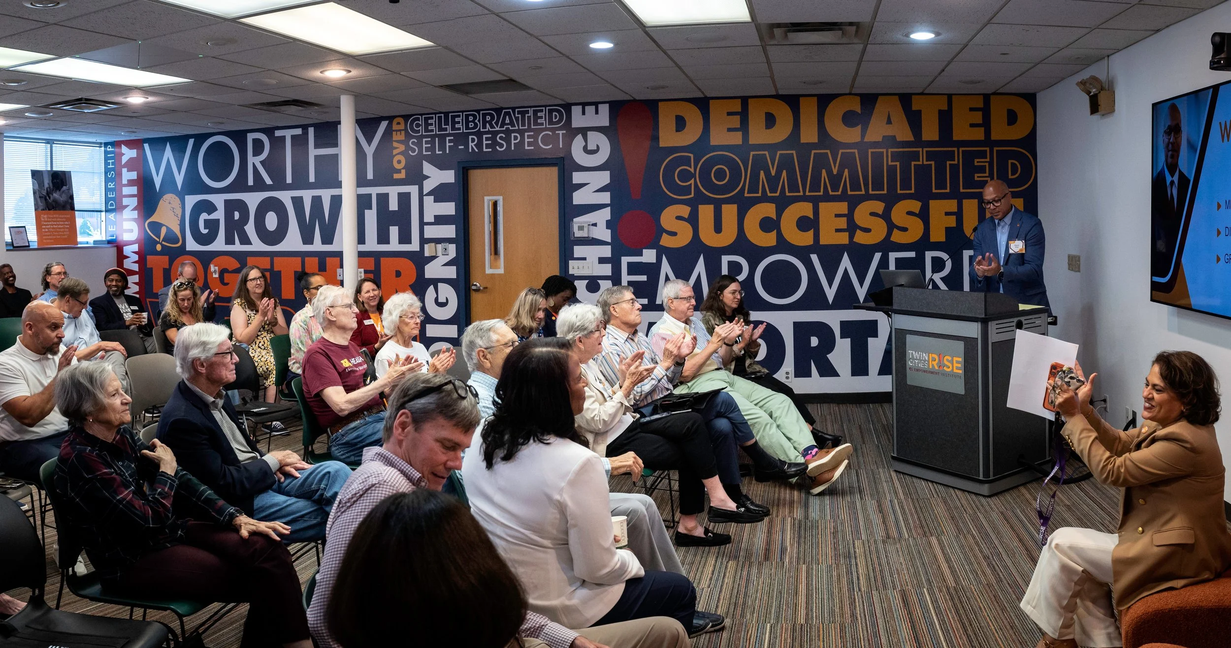 A group of diverse people sitting in chairs, some clapping, listening to a speaker at a podium labeled 'Twin Cities RISE.' The room has colorful wall art with words like 'worthy,' 'growth,' 'dedicated,' and 'successful.' A woman in the front is taking a photo of the speaker.