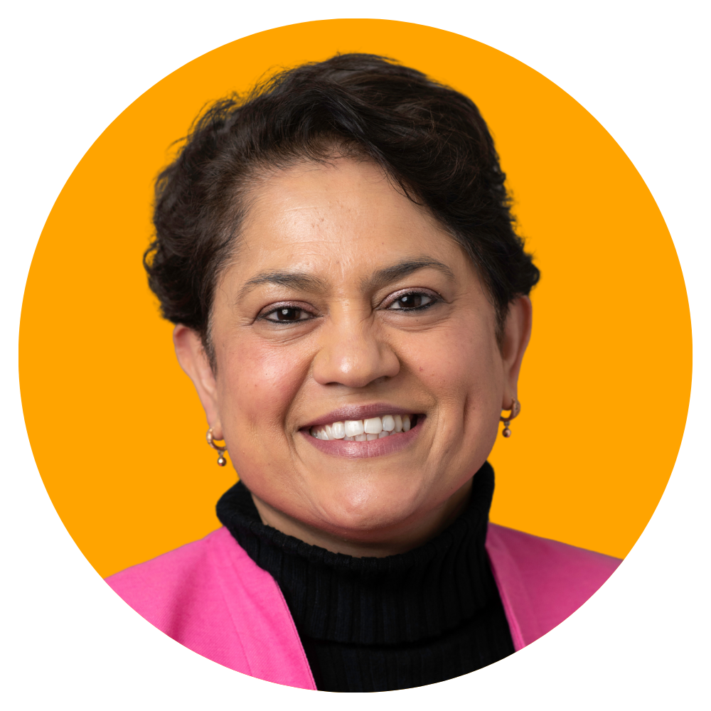 Close-up portrait of a woman with short dark hair, wearing a pink blazer over a black turtleneck, smiling, against an orange background.