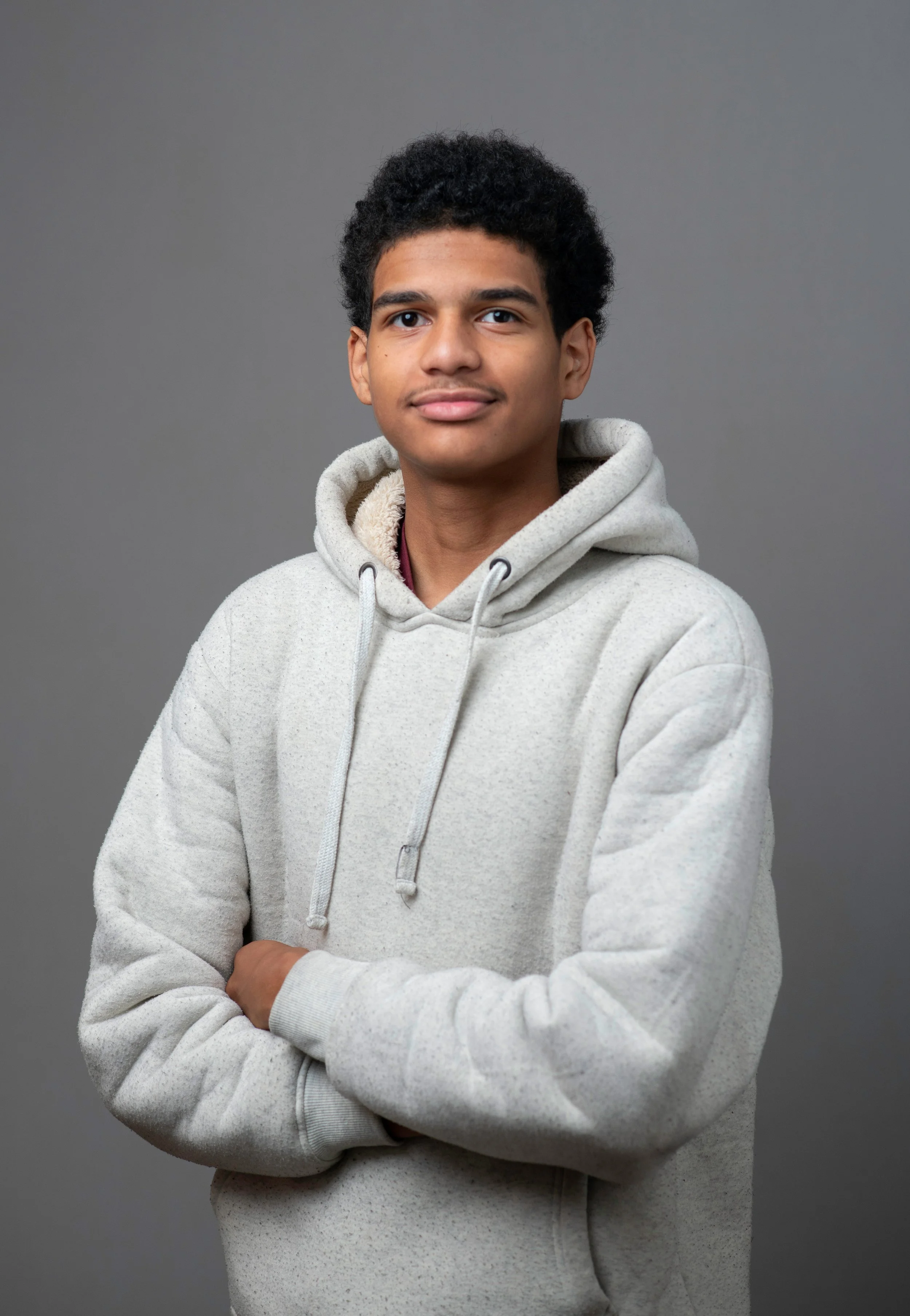 Young man with curly black hair wearing a light gray hoodie, standing against a gray background.