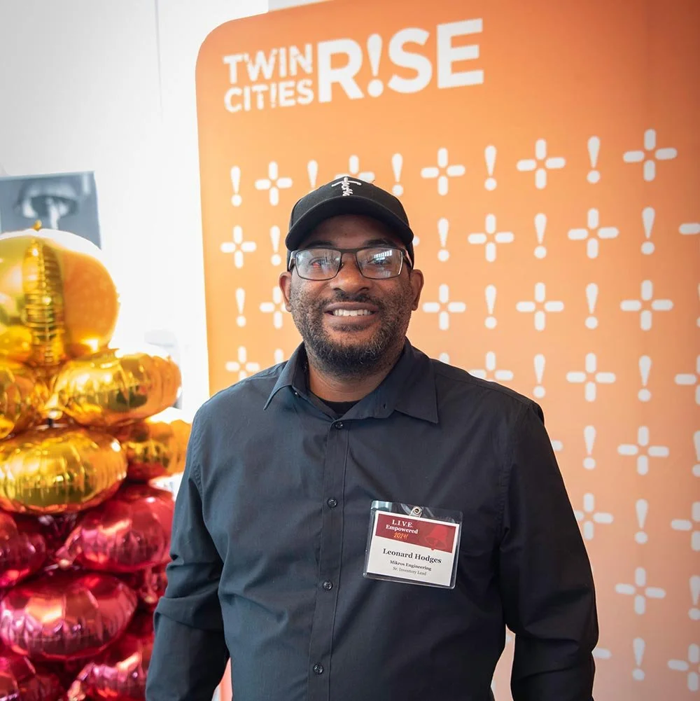 A man with glasses, a beard, and a black cap smiling at a conference or event. He is wearing a black shirt. Behind him is an orange banner with the words "Twin Cities RISE" and a design of white symbols, along with gold and pink balloons.