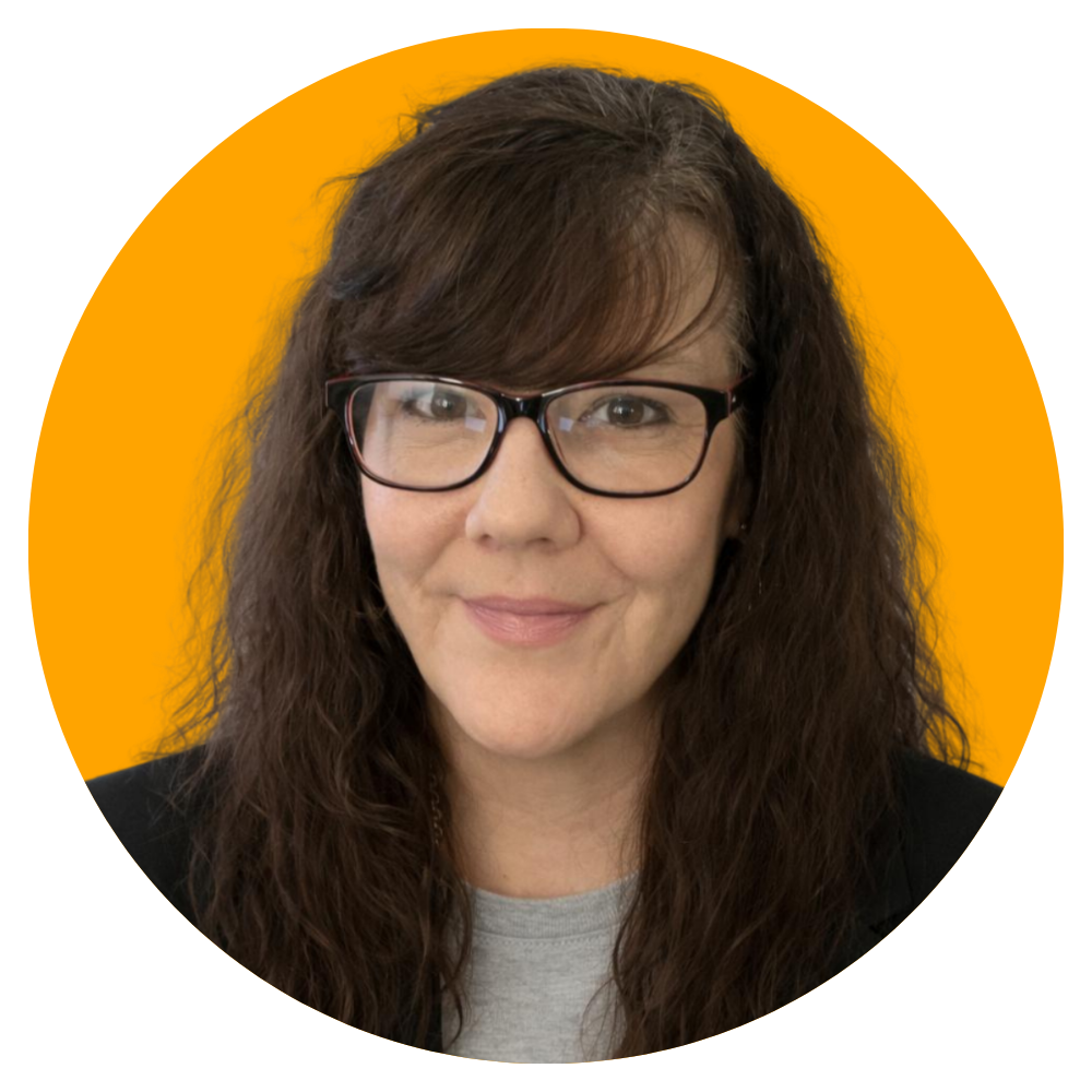 A woman with long brown curly hair, wearing glasses, a black blazer, and a gray shirt, smiling, with a solid orange background.