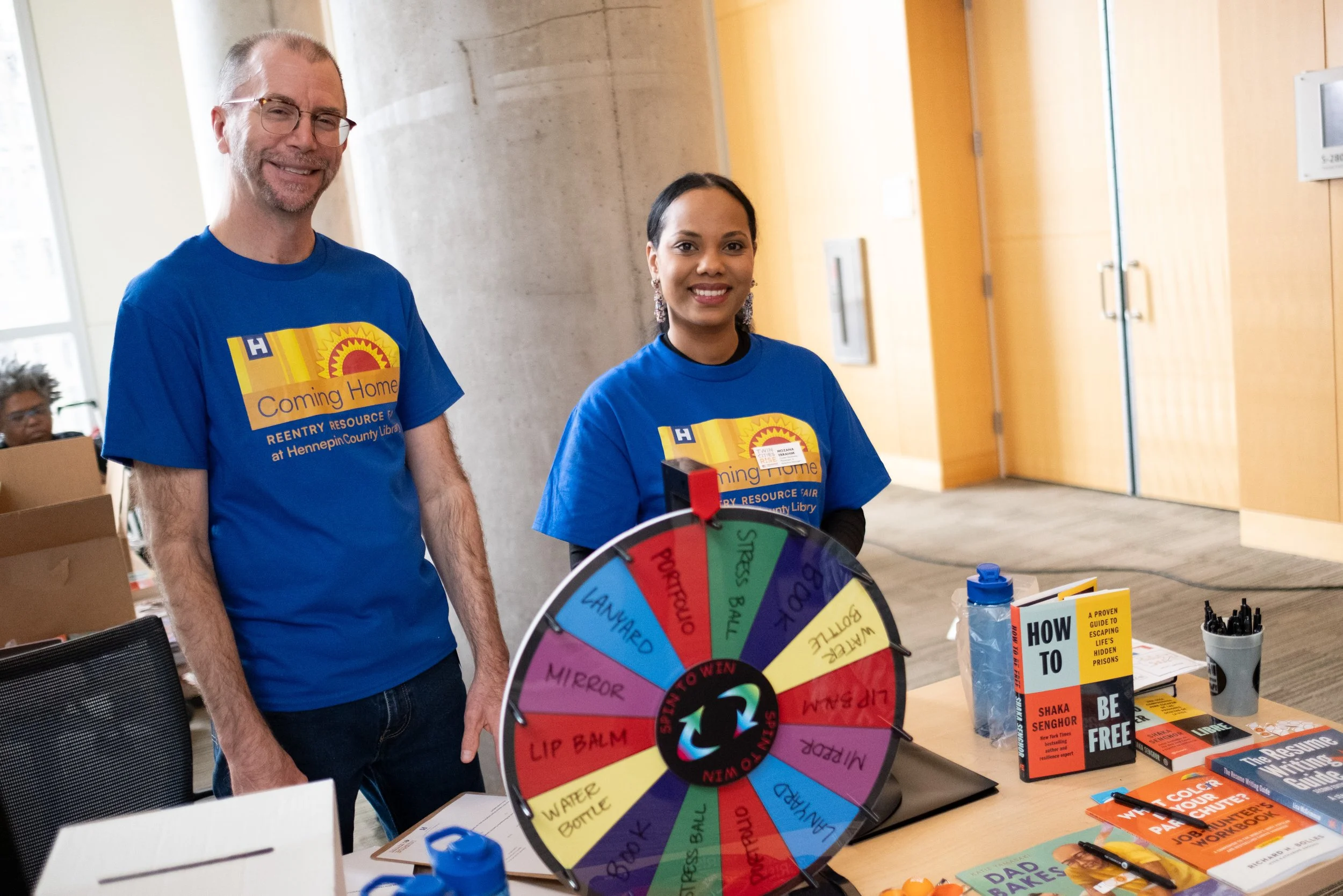 Two people, a man and a woman, wearing blue shirts with yellow and orange graphics, standing behind a table with a colorful spinning wheel labeled with various personal care items, at an indoor event for a library resource fair.