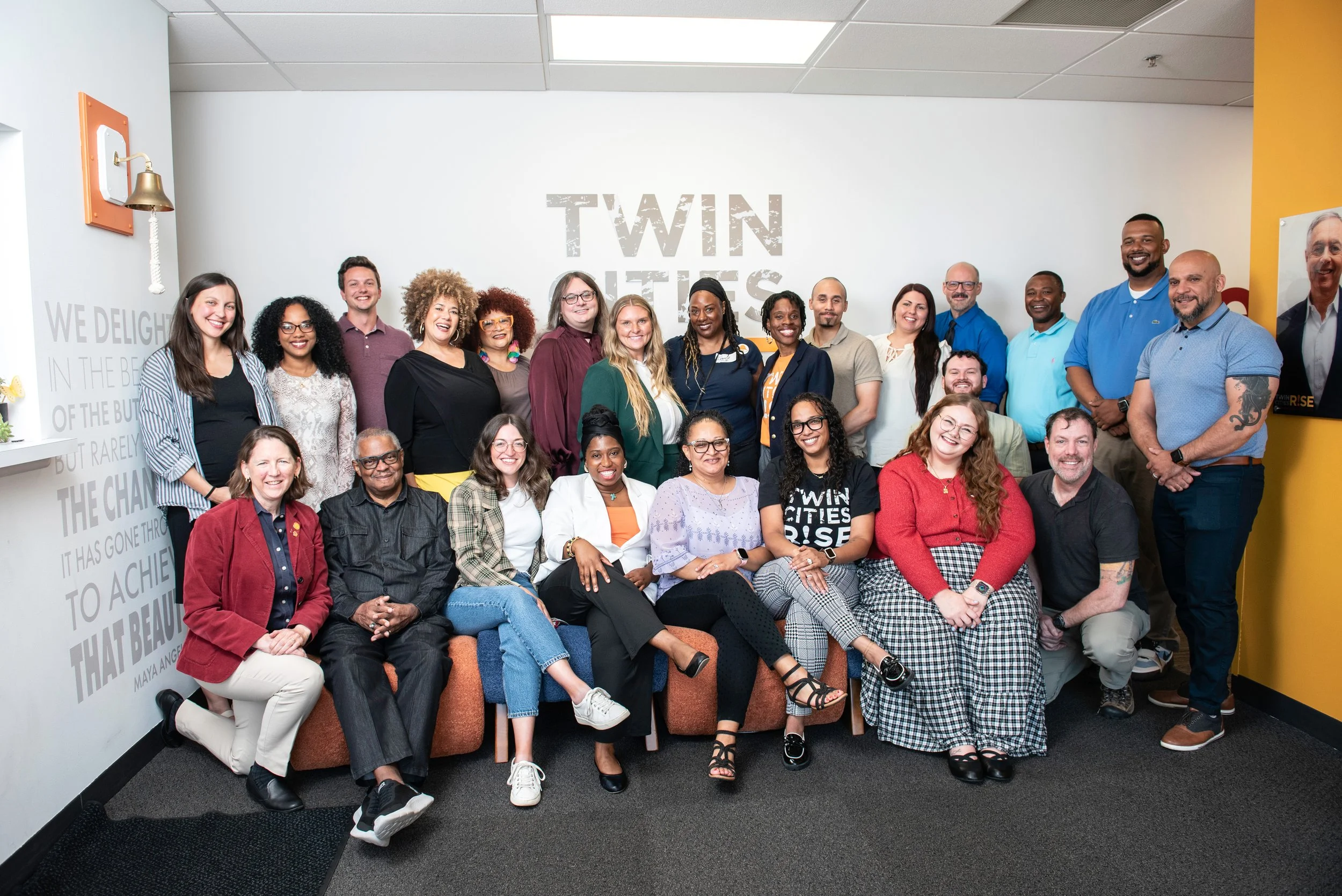 Group of diverse people smiling and posing for a photo in a room with a white wall that has the words 'TWIN CITIES RISE' written on it, some sitting on a couch and others standing.