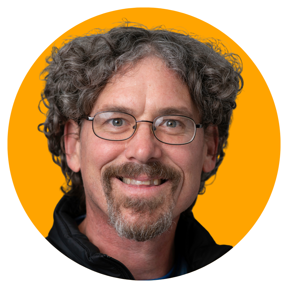 A middle-aged man with curly gray hair, glasses, and a mustache and beard, smiling against an orange background.