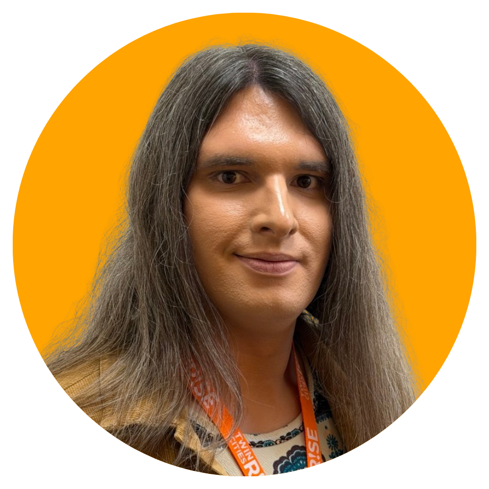 A person with long gray hair, light skin, and wearing a patterned shirt and orange lanyard posing against an orange background.