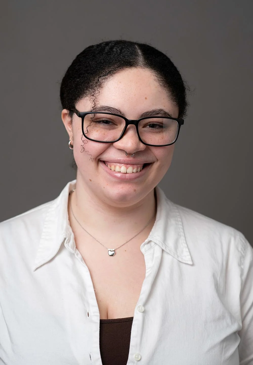 A woman with glasses and a septum piercing smiling, wearing a white button-up shirt and a necklace with a heart-shaped pendant, against a gray background.