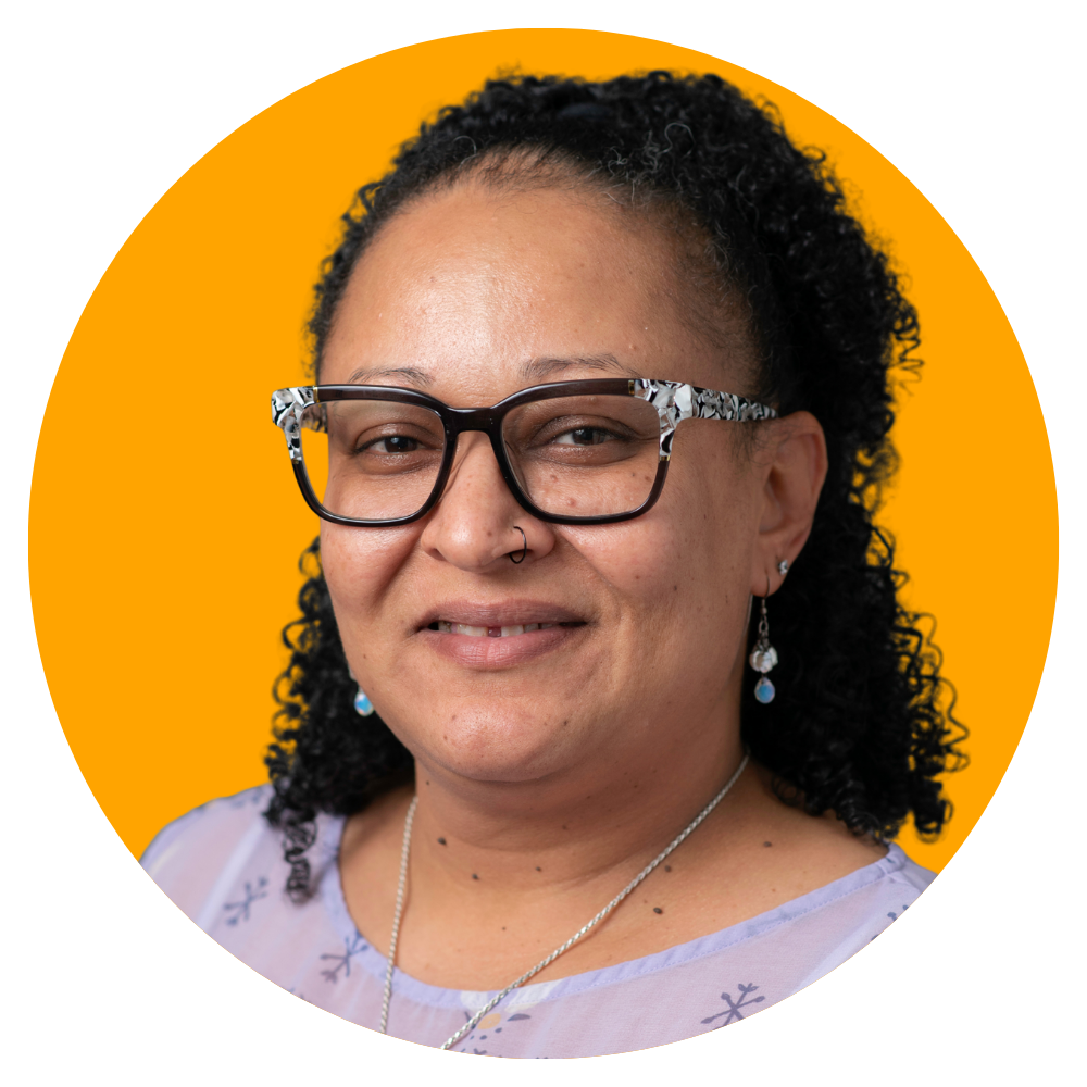 A woman with curly black hair wearing large black and white glasses, earrings, a nose ring, and a necklace, smiling against a yellow background.