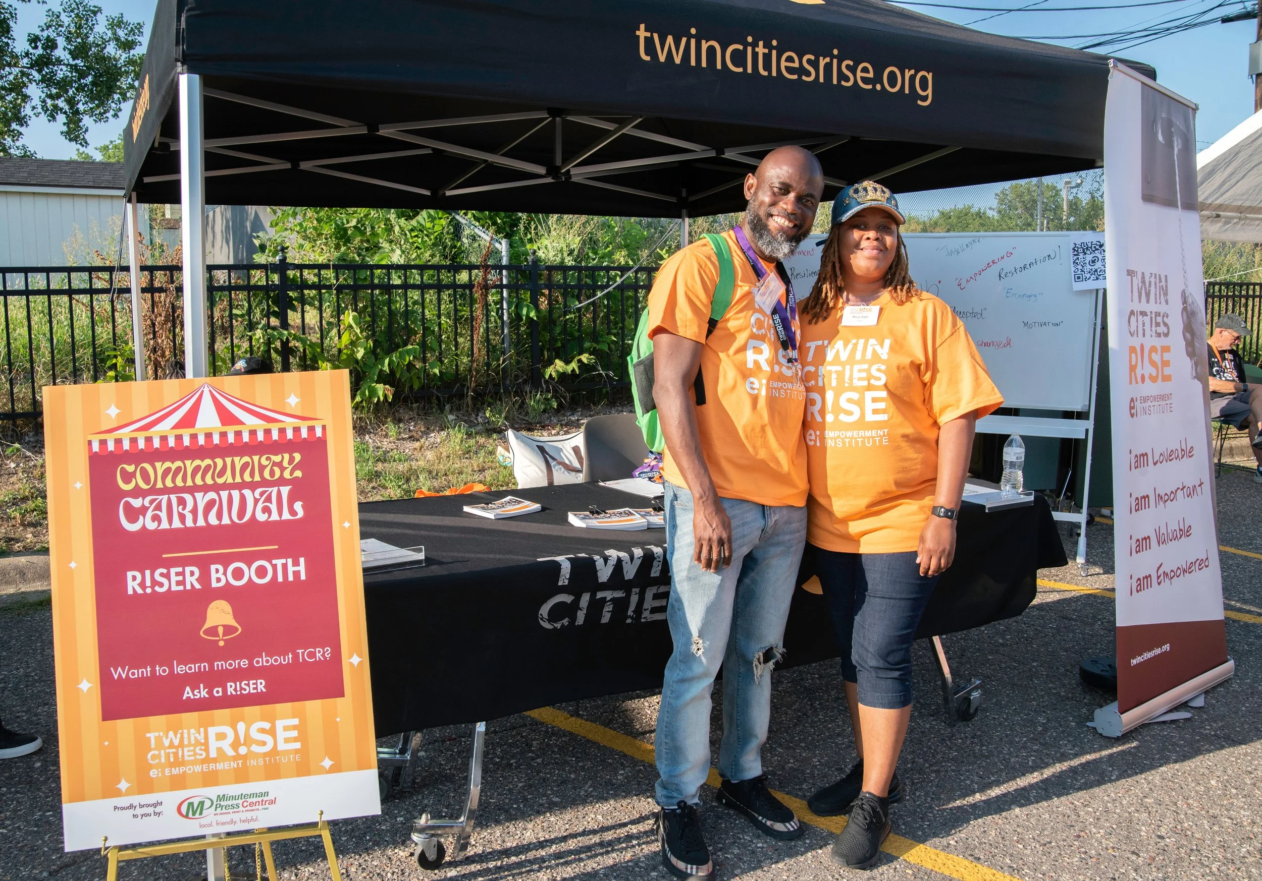 Two people smiling at an outdoor community event stand under a black canopy with the website 'twincitiesrise.org' printed on it. They are wearing orange t-shirts with the text 'TWIN CITIES RISE'.
