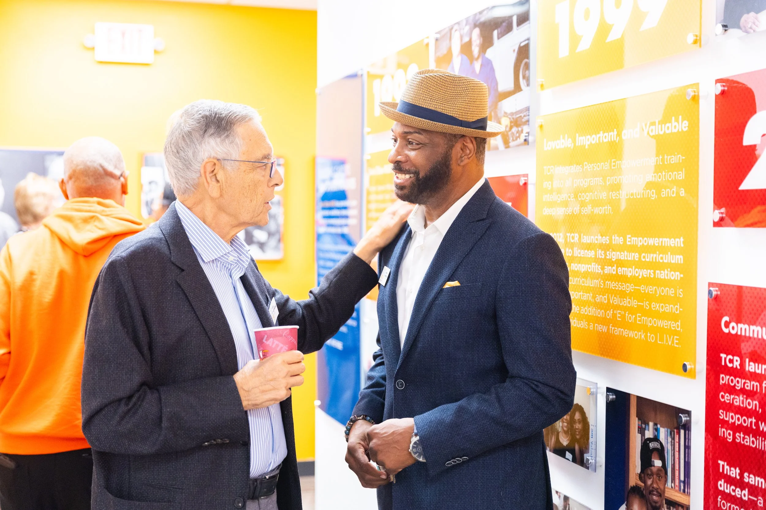 Two men are engaged in conversation at an indoor event or exhibition, standing near display boards with informational posters. One man has gray hair, glasses, and a business suit, while the other man has a beard, a straw hat, and a dark blazer.