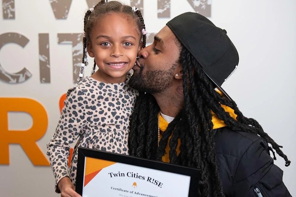 A young girl with braided hair smiling while holding a certificate. A man with long dreadlocks, a cap, and a yellow hoodie is kissing her on the cheek, standing behind her.