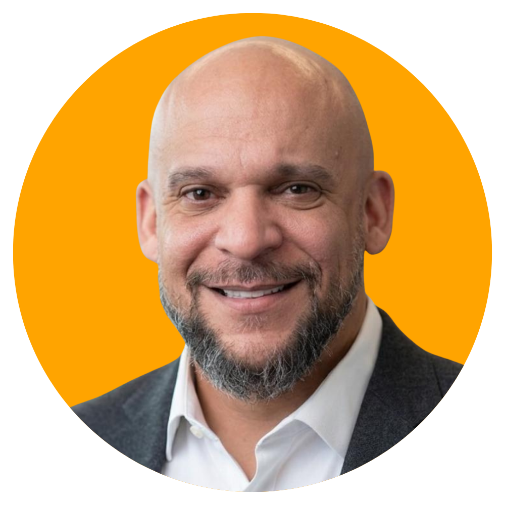 A smiling bald man with a beard, wearing a white shirt and gray blazer, against an orange background.