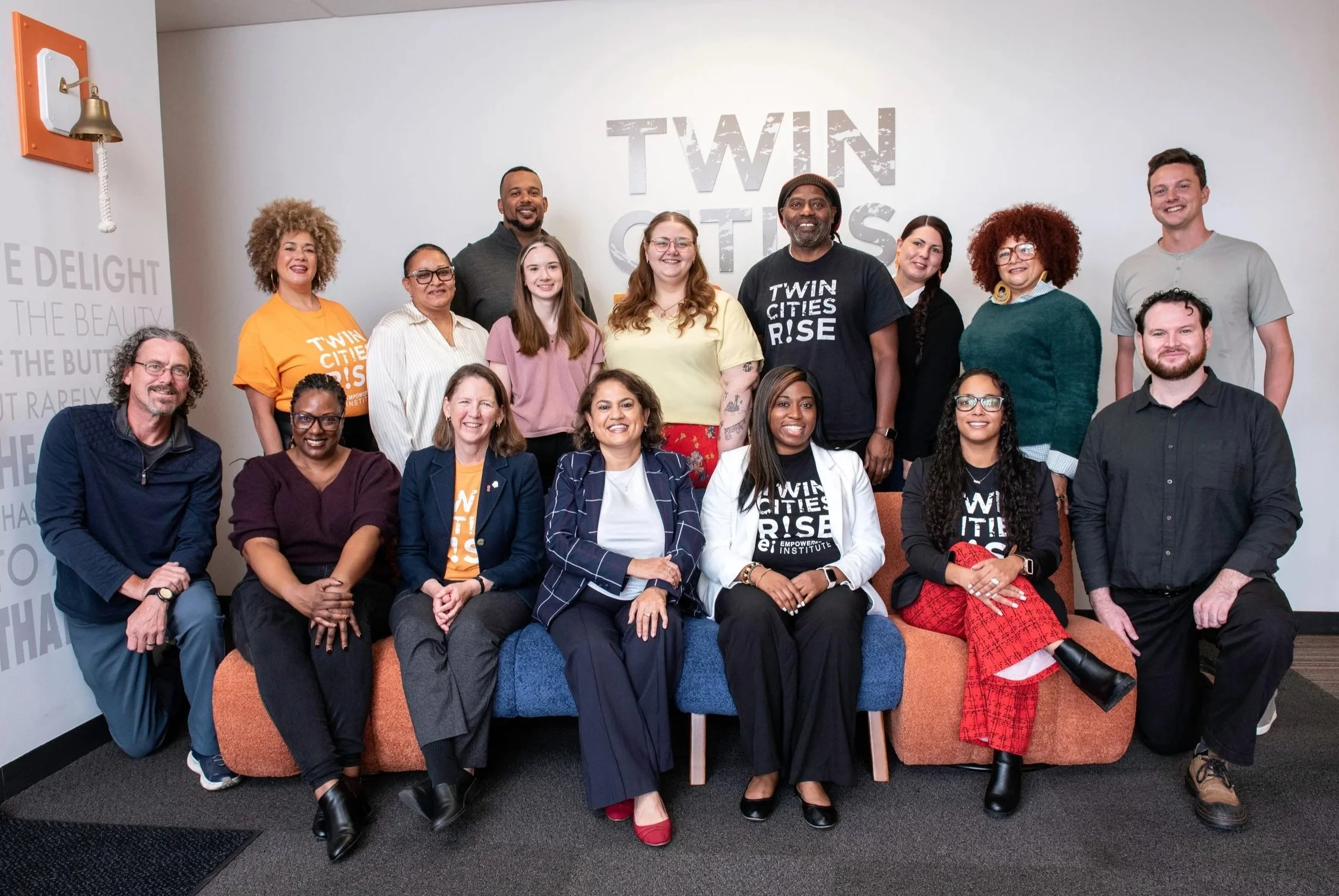 A diverse group of 16 people posing together for a photo in an indoor setting, some sitting on a couch and others standing behind. The background features a wall with the words 'TWIN CITIES RISE' and a partial quote on the side wall. The group includes men and women of different ages and ethnicities, dressed in casual and business casual attire.
