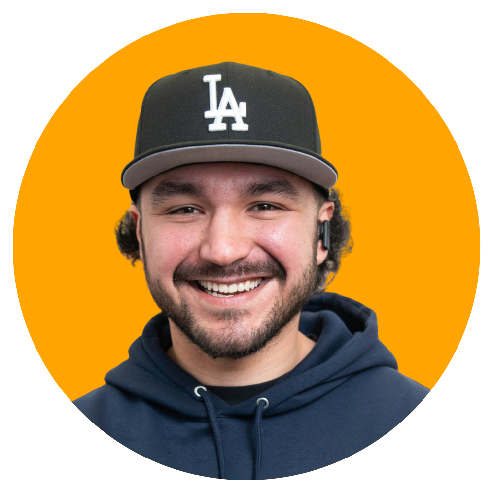 A smiling man wearing a black LA Dodgers cap, a dark hoodie, and a wireless earpiece against an orange background.