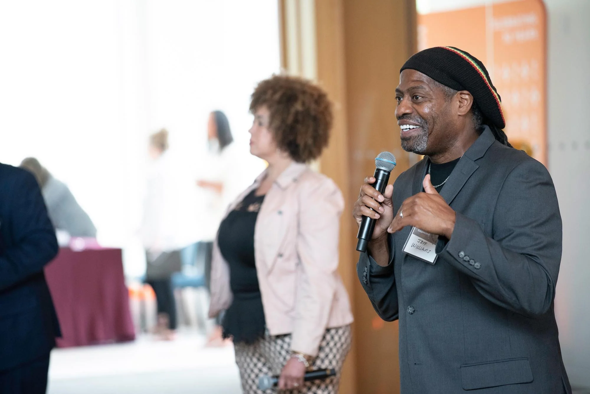 Man wearing a black hat, dressed in a gray suit, speaking into a microphone during an event. In the background, there is a women with curly hair standing near a table with a burgundy tablecloth.